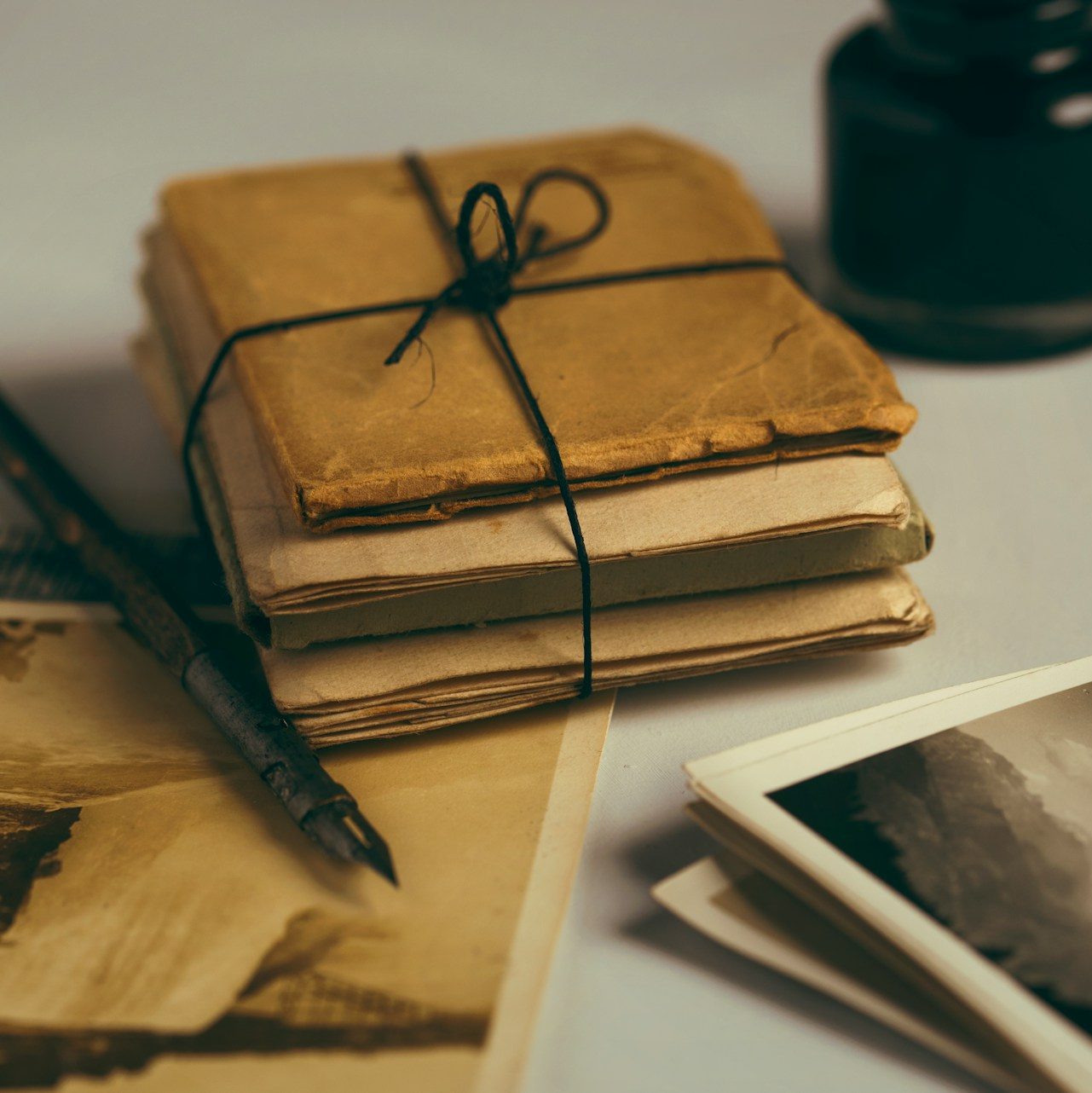 Two stacks of old, yellowed photos with a stack of brown paper letters tied together with brown string. Next to the letters is a fountain pen and a bottle of black ink.