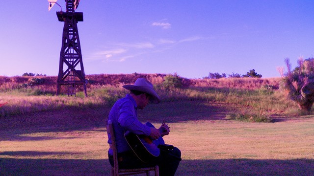 Man in cowboy hat sitting, playing guitar. Background, desert with mesa, sun setting, windmill